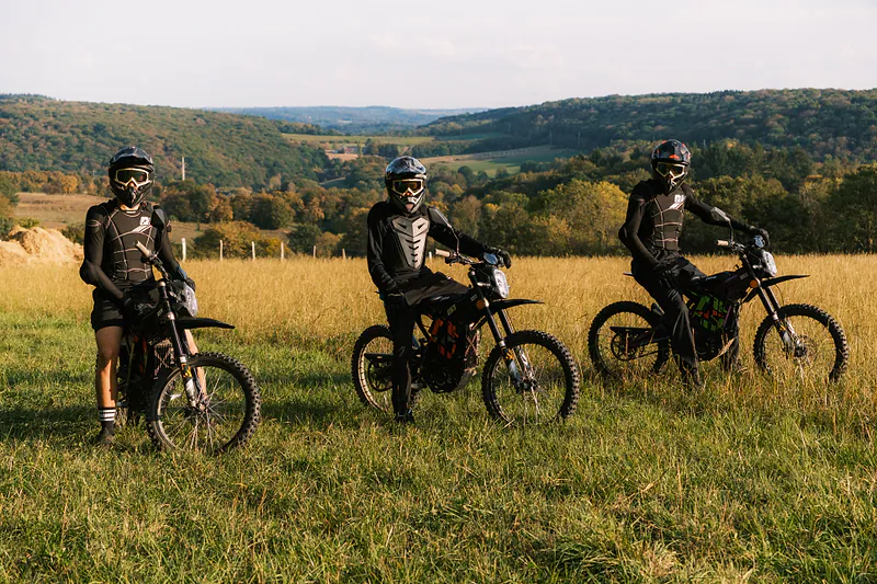 Trois pilotes en Sur-Ron dans un champ doré, avec la vallée et les bois en arrière-plan