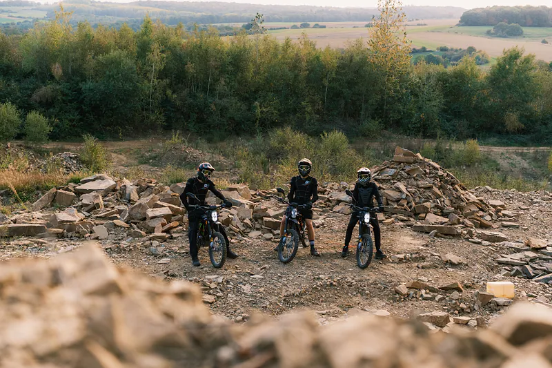 Trois pilotes alignés sur leurs motos au milieu d’un terrain de pierres