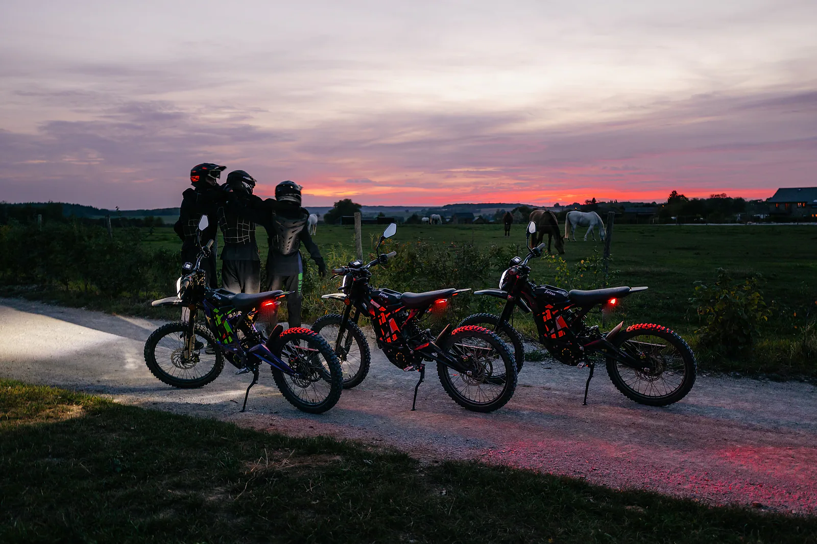 Trois motos électriques Sur-Ron, feux allumés, sur un chemin du Pré Mathy au coucher du soleil, avec leurs pilotes et des chevaux dans le pré